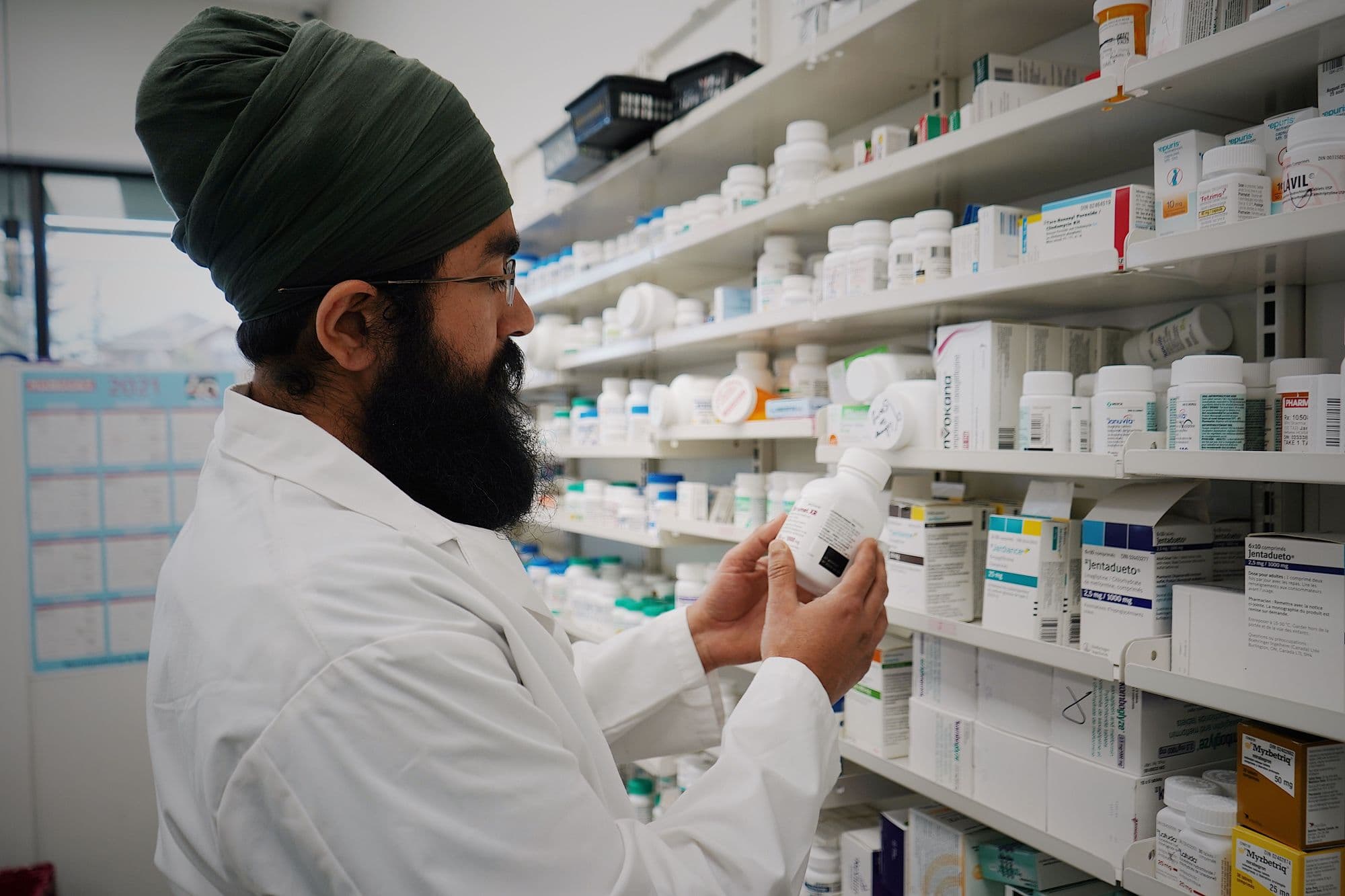 A pharmacist in a white coat and turban reviews a medication bottle in front of stocked pharmacy shelves.