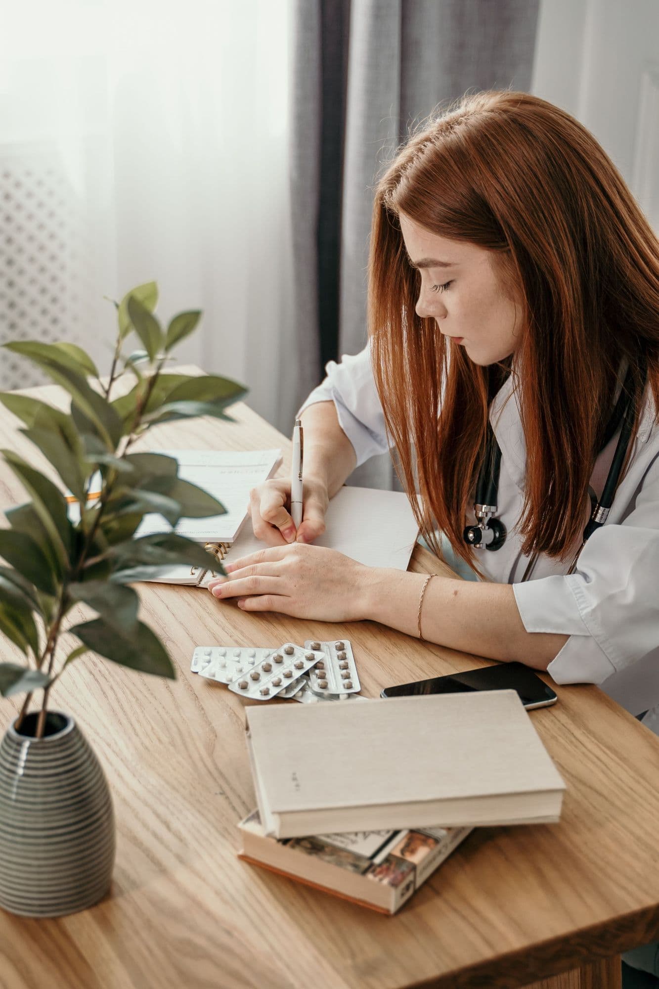A pharmacy student studies at a wooden desk with notes, textbooks and a plant.