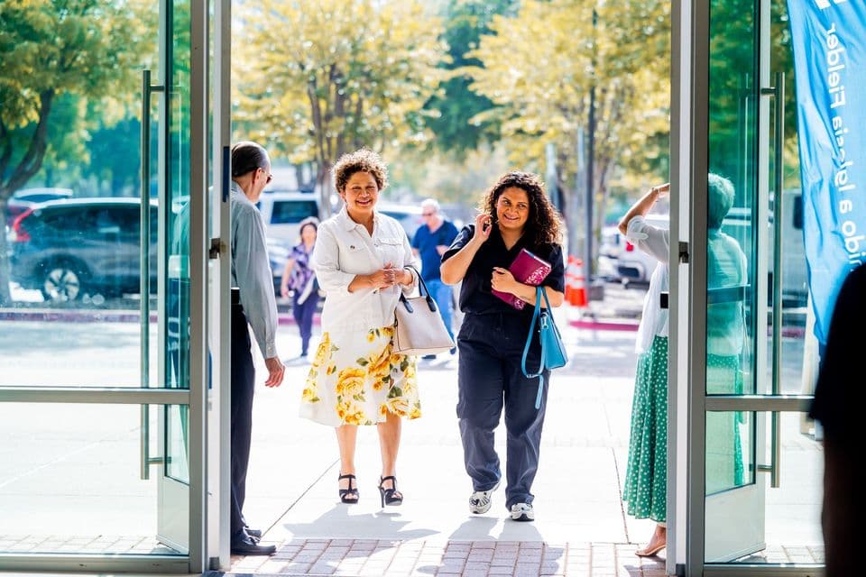 Two smiling women walk through open glass doors into a community event on a sunny day.