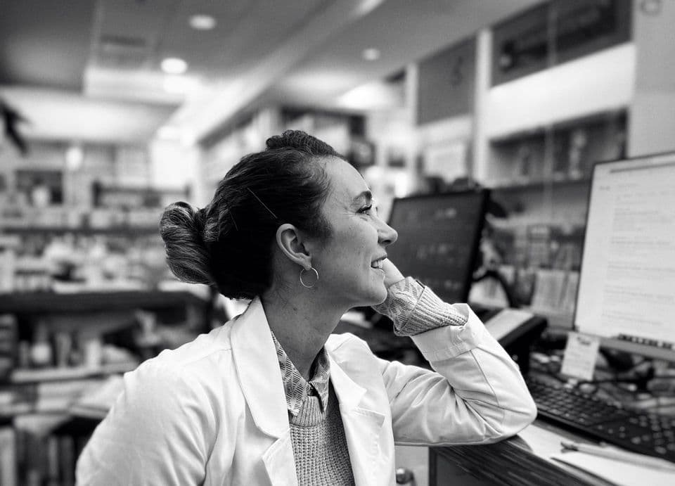 Black-and-white photo of a smiling pharmacist leaning on the dispensary counter.